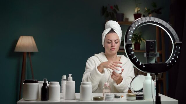 Female beauty blogger wearing bathrobe showing face cream recording video for social media