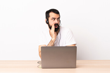Telemarketer caucasian man working with a headset and with laptop whispering something with surprise gesture while looking to the side.