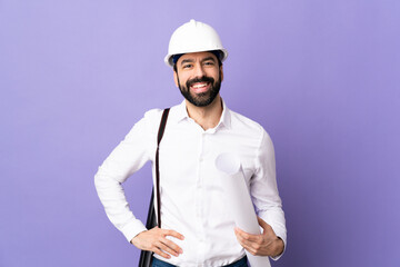 Young architect man with helmet and holding blueprints over isolated purple background posing with arms at hip and smiling