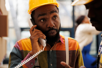 African american man talking on landline phone in industrial storehouse. Factory warehouse operator answering supervisor telephone call and listening to inventory management instructions