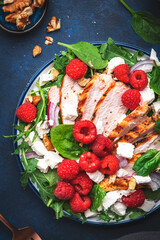 Tasty salad with raspberries, grilled chicken, cheese, onion, walnuts, spinach and arugula, blue table background, top view