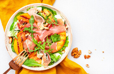 Tasty salad with grilled peach, jamon, feta cheese, walnuts and arugula on white table background, top view