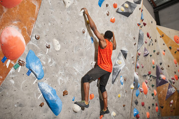 athletic african american man in orange shirt gripping on boulders while climbing up rock wall