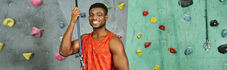 joyful athletic african american man in orange shirt smiling happily at camera, bouldering, banner