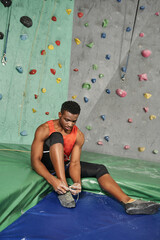 vertical shot of sporty african american man on crash pad tying his shoes with rock wall on backdrop