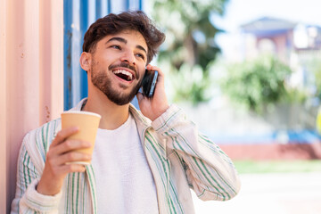 Handsome Arab man at outdoors using mobile phone and holding a coffee with happy expression