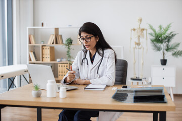 Focused female doctor with stethoscope having video conference on modern laptop. Attractive hindu professional in doctors coat explaining important things during online conversation with patient.