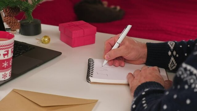 Writing a Christmas Wishing Card. Male adult making greeting cards at home office during Xmas. Man hands in blue sweater holding blank paper and envelope at the Desktop. Coffee Cup, holiday decoration