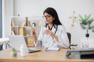 Attentive indian woman holding medicines and telling about it effects during video call on wireless laptop. General practitioner in medical uniform having online appointment with sick patient.