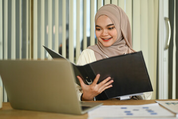 Fototapeta premium Smiling Muslim businesswoman reading document, checking financial report at office desk
