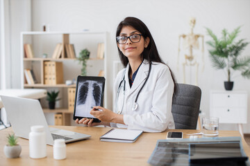 Concentrated female doctor talking on video call while showing result of examination of patient with tuberculosis. Indian lady consulting person using laptop and tablet in spacious clinic room.