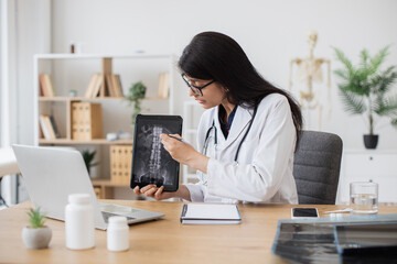 Indian female specialist sitting in front of digital laptop and consulting patient with spinal...