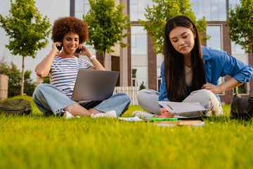 Two female students doing homework together while sitting on lawn in campus