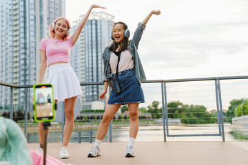 Two joyful girls making dancing videos with smartphone camera on city waterfront