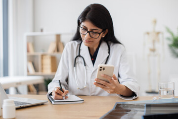 Attentive indian doctor sitting at table with smartphone and writing data of new patient into...