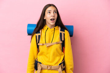 Young mountaineer girl with a big backpack isolated on pink background looking up and with surprised expression