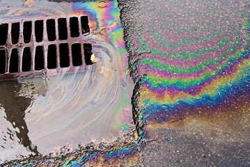 An oil slick against the backdrop of an asphalt road flows into a storm drain through a grate.