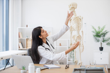Qualified female medic with stethoscope analyzing skeleton anatomical model at cozy workplace....