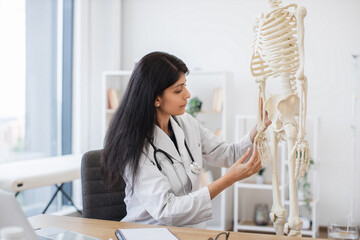 Competent female physician examining hands bones on plastic skeleton while sitting at workplace in...