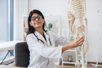 Portrait of positive hospital worker in eyeglasses and lab coat sitting next to model skeleton and...