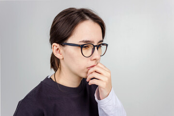 Young brunette girl student with glasses looks down thoughtfully. She is confused. Isolated on grey background.