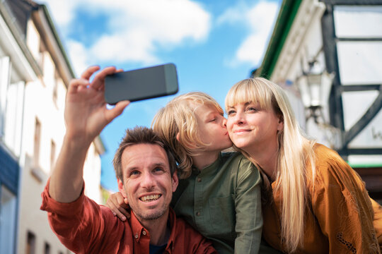 Smiling father taking selfie with son kissing mother on sunny day
