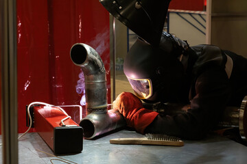Welder at work. Worker connects two curved pipes. Man wearing soldering mask. Welder works at table for soldering metal. Manufacturing of complex pipeline section. Welder works in factory or plant