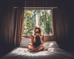 mulher sentada em cama com janela aberta ao fundo tomando uma xícara de café 