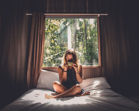 mulher sentada em cama com janela aberta ao fundo tomando uma x&iacute;cara de caf&eacute; 