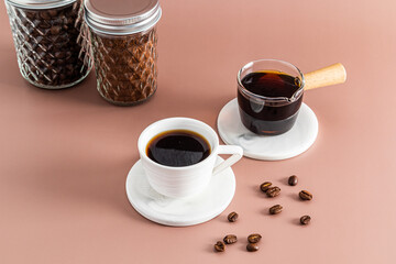 A small white cup of freshly brewed espresso coffee and a glass cezve with coffee on a beige background with two glass jars of ground coffee.