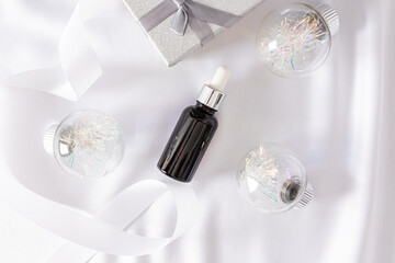 Cosmetic bottle with dropper and silver cap lies on white satin among silver Christmas balls and gift box. Top view. Empty product packaging.
