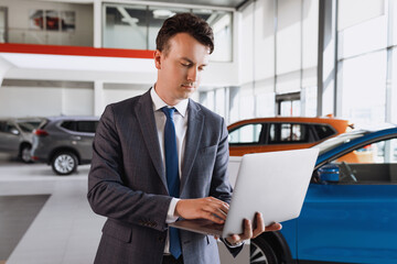 Portrait of a successful head of a dealership in a suit who works on a laptop against the background of cars.