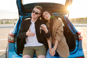 A young couple is sitting in the trunk of their new car. The man holds the key. Against the background of sunset.