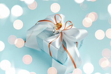 Gift box wrapped in blue silver paper with white and gold ribbon bow. Blue background, bokeh light. Christmas and New Year gifts, Boxing Day.