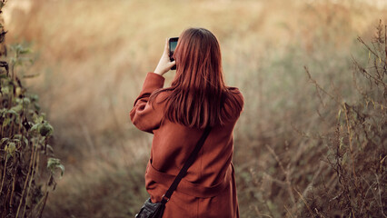 Back of girl with long red hair in red coat, standing among autumn grasses and taking photo of landscape on a smartphone.