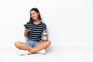 Young caucasian woman sitting on the floor isolated on white background holding coffee to take away and a mobile