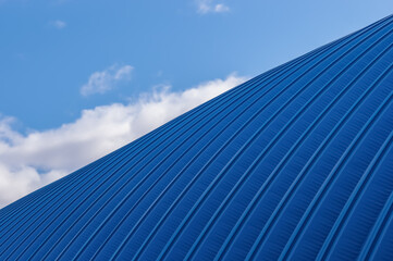 Fototapeta na wymiar A fragment of a hangar made of blue rolled metal against the background of the sky with clouds. A simple metal structure of an industrial building from the outside.