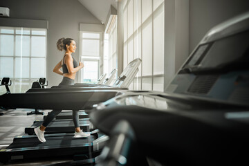 Full length shot of Beautiful woman with fit body in sportswear running on a treadmill near panoramic window in modern gym. Sun shining through window - obrazy, fototapety, plakaty