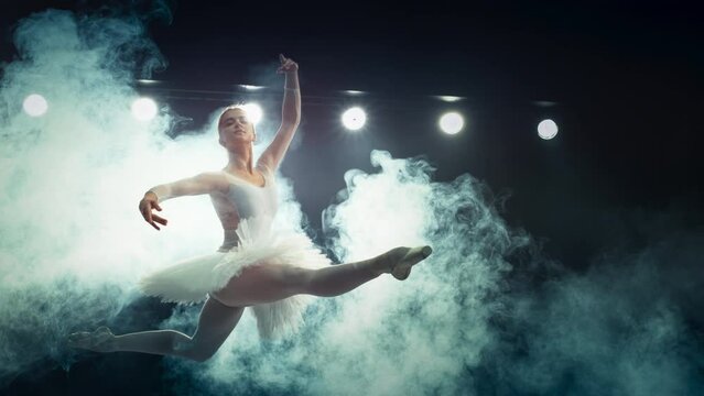 Modern Ballet Performance on a Big Stage with Lights. Beautiful Female Ballerina in White Dress Making a Perfect Split Leap Through the Smoke. Super Slow Motion Speed Ramp Effect