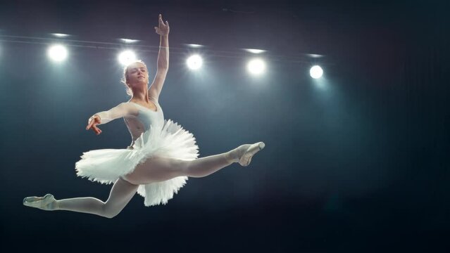 Graceful Ballet Dancer Performs a Powerful Split Leap During Her Act. Young Female Practicing in Striking Beautiful Dress in a Dark Room. Cinematic Super Slow Motion Speed Ramp Footage