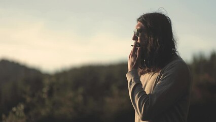 A man with a beard with long hair, wearing glasses and headphones, smoking a cigarette and looking at a beautiful sunset in the mountains. Trekking and hiking in the forest, the concept of success
