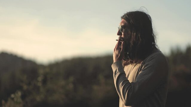 A man with a beard with long hair, wearing glasses and headphones, smoking a cigarette and looking at a beautiful sunset in the mountains. Trekking and hiking in the forest, the concept of success