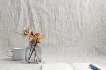 A transparent vase with orange-tinged roses and other dried flowers in front of a white mug against a light gray crinkled fabric background. In the foreground is a white notebook with a blue pen on to