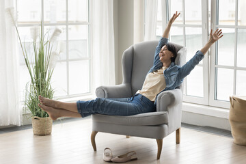 In cozy living room excited middle-aged woman enjoy relaxation seated on armchair with arms raised...