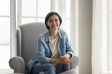 Middle-aged pretty woman in denim shirt and jeans posing for camera seated on modern armchair,...