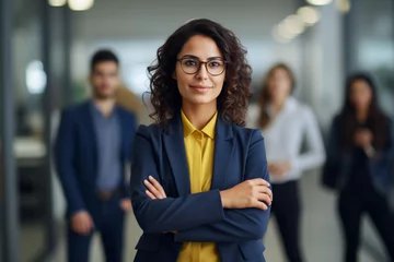 Foto op Canvas Portrait of one confident young hispanic business woman standing with arms crossed in an office with her colleagues in the background, Ambitious entrepreneur and determined leader ready for success  © alisaaa