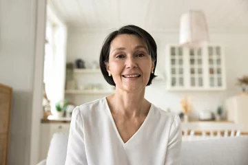 Fotobehang Attractive middle-aged woman smile looks at camera sit on sofa, having videoconference remote talk to family or grown-up children living abroad, enjoy communication using modern tech, profile picture © fizkes