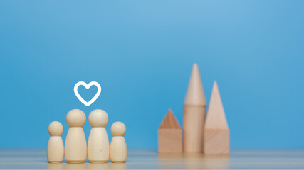 Housing for families. Close up of wooden figurines of family with children and model of small one-story house. Miniature figurines made of light wood stand on table in bright room. Blurred background.