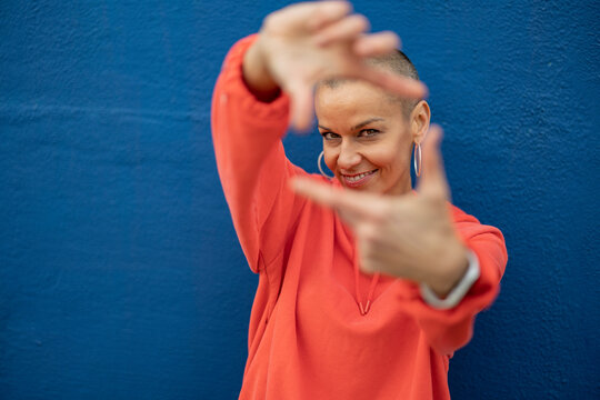Smiling woman with shaved head showing finger frame in front of blue wall