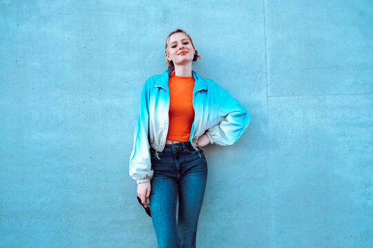 Smiling young woman with hand on hip standing in front of blue wall